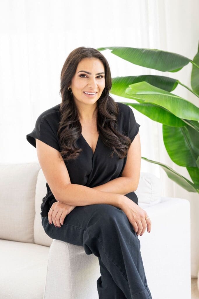 woman sitting with legs crossed on arm of couch smiling at camera. wearing all black. 