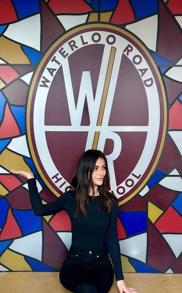 Carla Cressy sat with her arm raised in front of 'Waterloo Road High School' wall