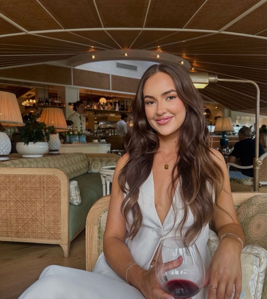 Woman with long brown hair holding a glass of red wine, sat in a chair legs crossed smiling at camera