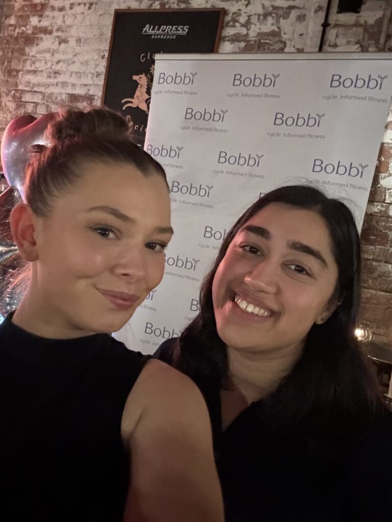 A selfie of two women smiling in front of a white Bobbi Cycle Informed Fitness brand banner at an indoor event