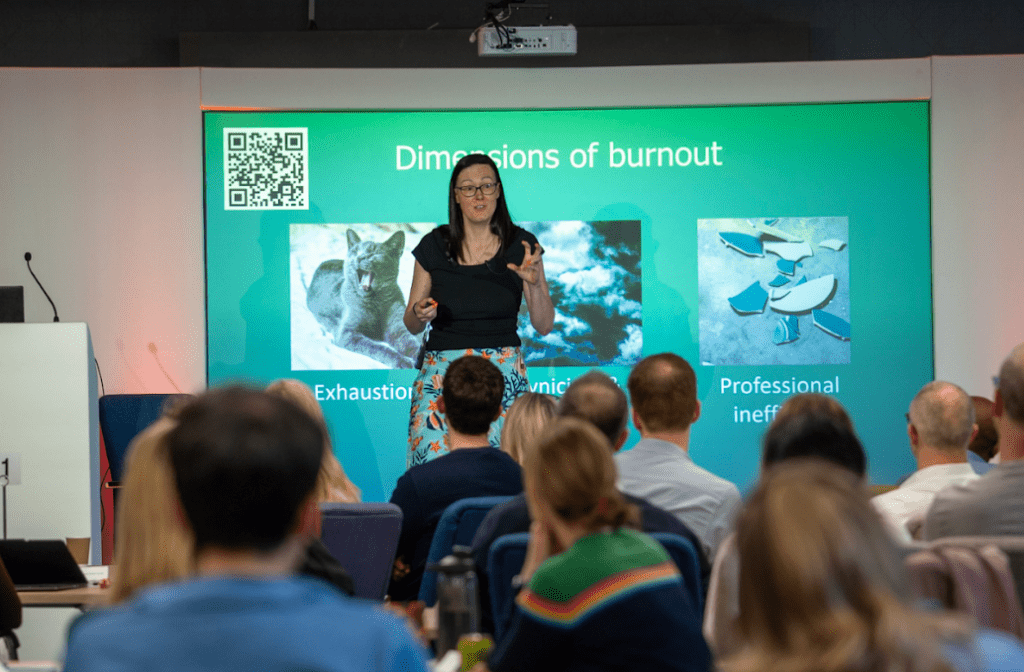 Woman wearing a black shirt and blue skirt presenting to an audience - background is a bright blue slide
