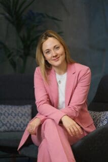 Woman with blonde hair wearing pink suit, seated with legs crossed smiling at camera 