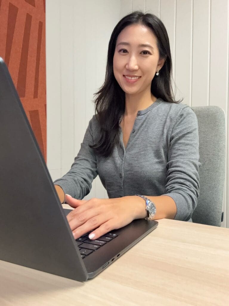 Woman with dark brown/black hair smiling at camera, sat at desk, typing on computer 