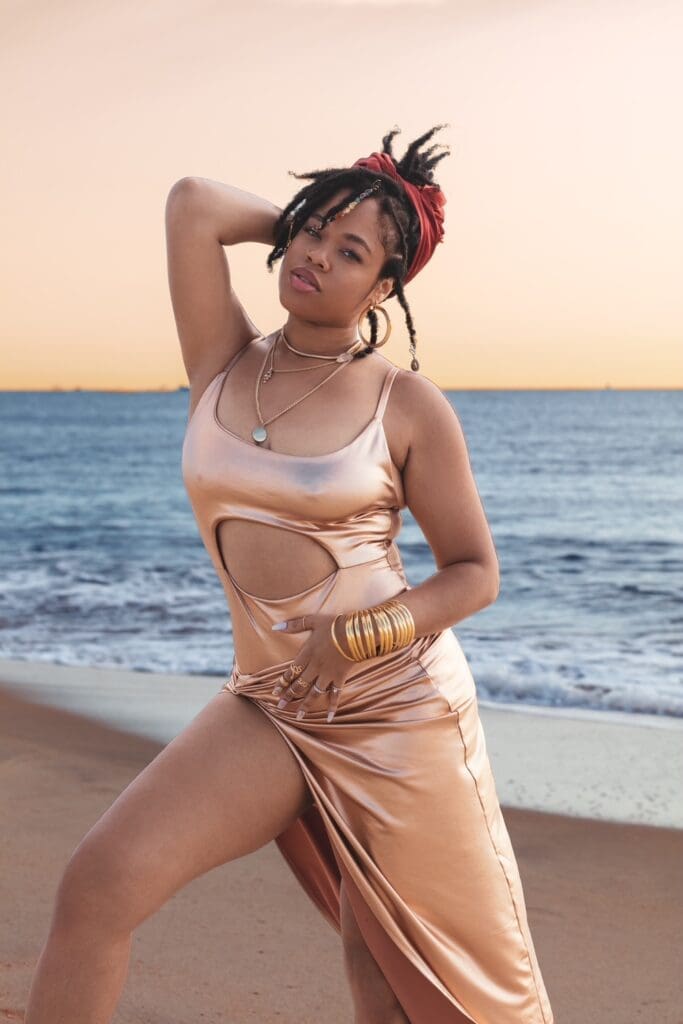 Girl posing on beach - wearing a pink dress with one hand on her stomach and the other behind her head.