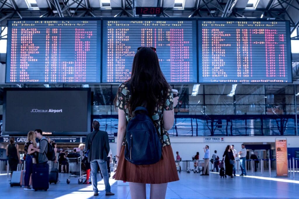 Woman in an airport with her back to the camera, standing staring at the Flight Information Display System carrying a backpack