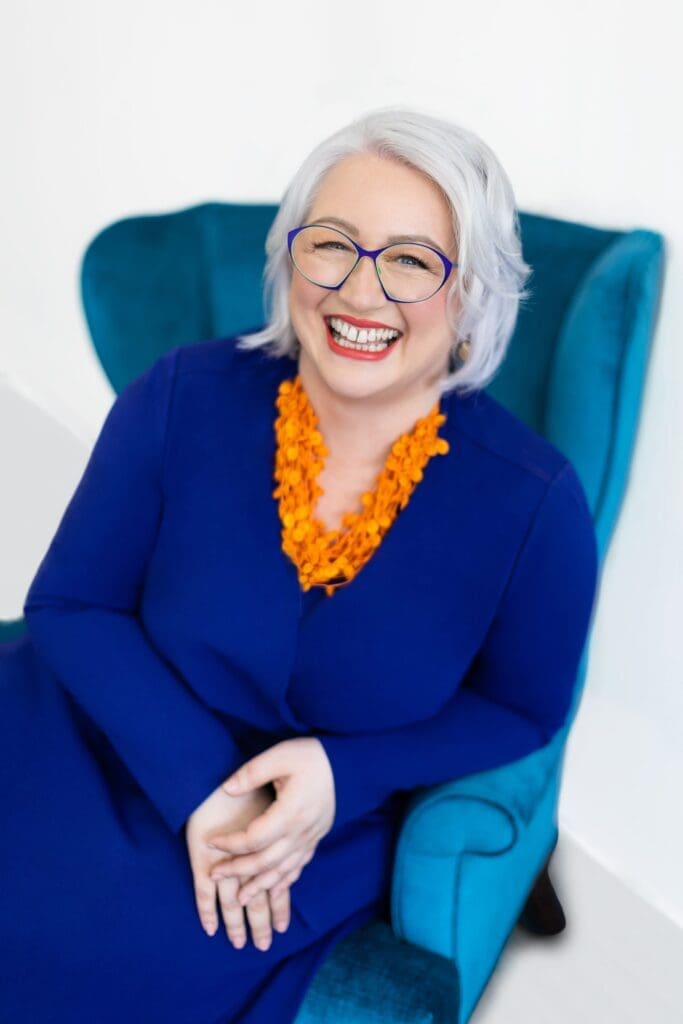 Woman sat in chair smiling wearing a royal blue dress and bright orange necklace 