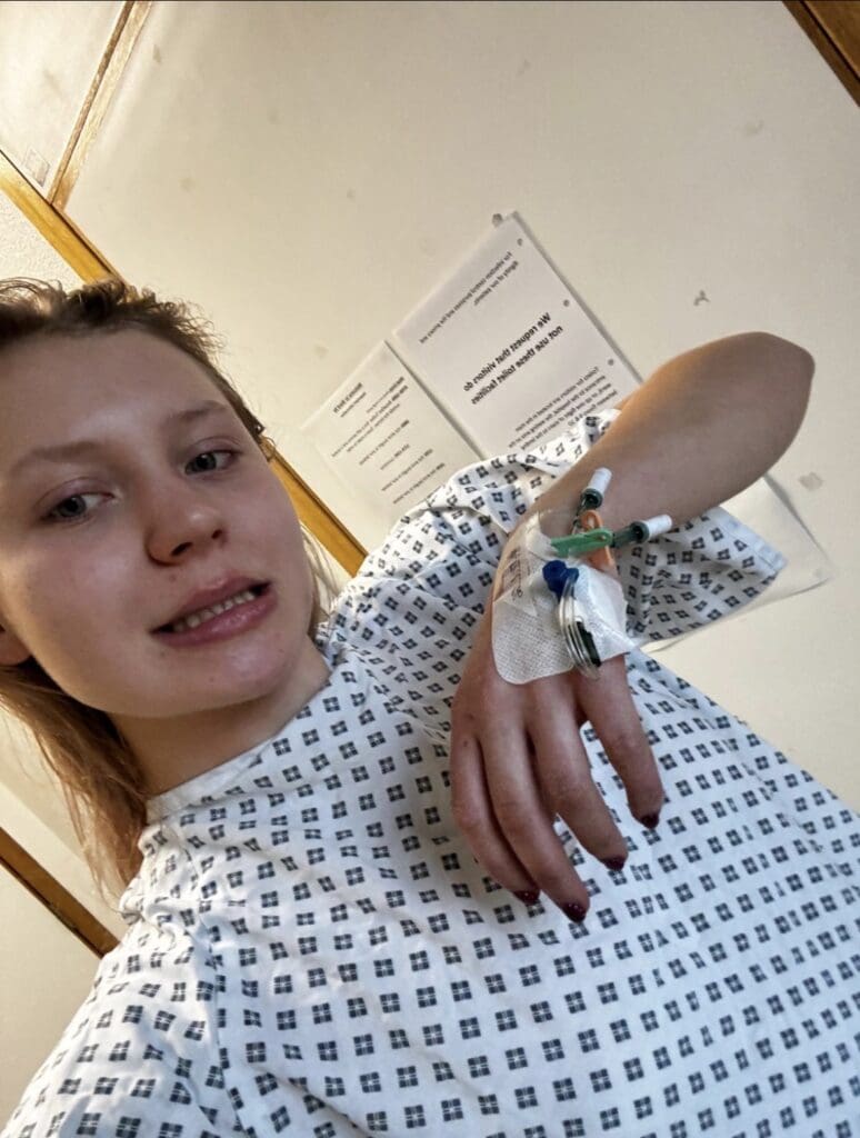 Woman in hospital gown showing IV in her hand to camera 